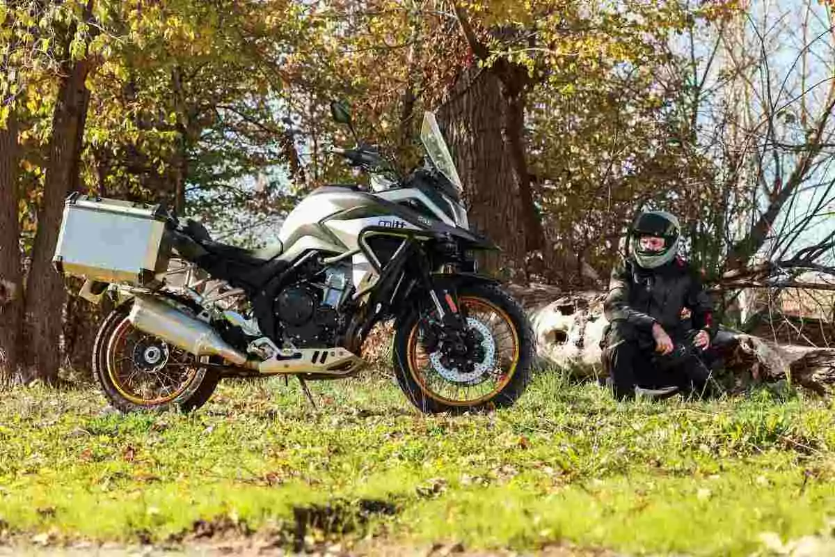 Motocicleta de aventura estacionada en un área boscosa con una persona usando equipo de protección sentada en el suelo junto a un tronco caído Motocicleta de aventura estacionada en un área boscosa con una persona usando equipo de protección sentada en el suelo junto a un tronco caído