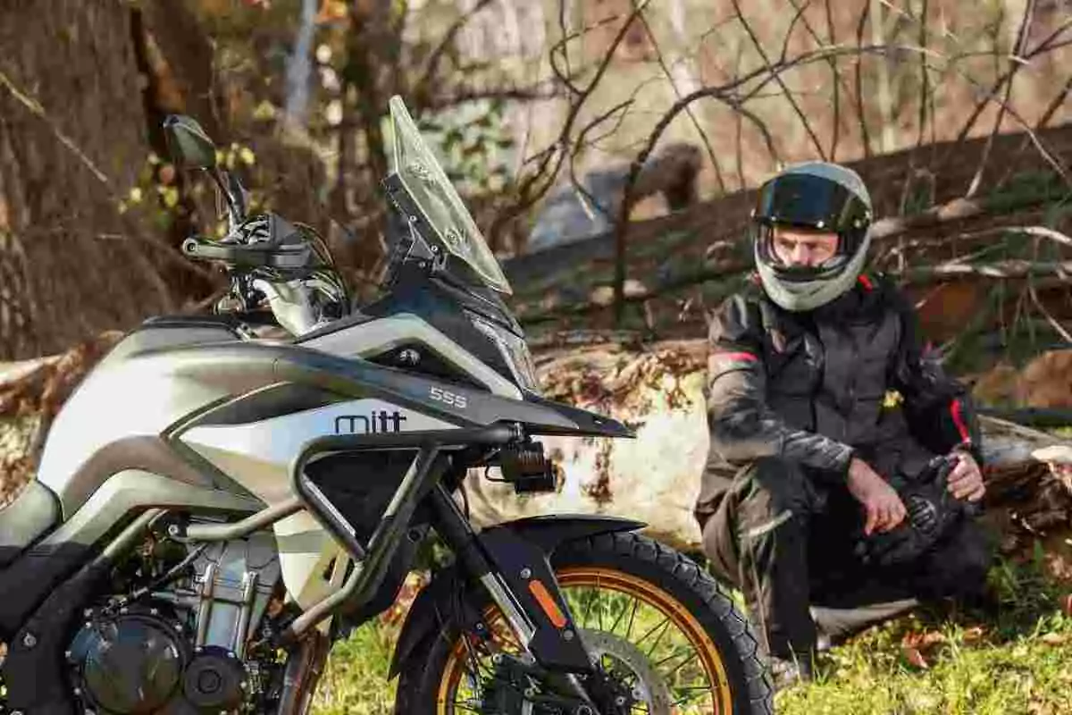 Motocicleta de aventura estacionada en un entorno natural con un hombre usando casco y ropa de protección sentado en el suelo junto a un tronco caído Motocicleta de aventura estacionada en un entorno natural con un hombre usando casco y ropa de protección sentado en el suelo junto a un tronco caído