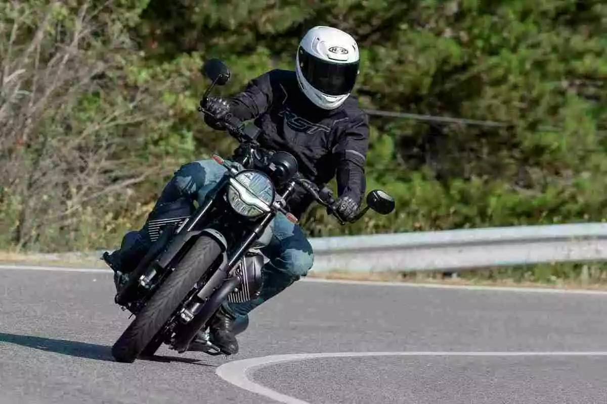 Persona con casco blanco y chaqueta negra conduciendo una motocicleta en una curva de carretera rodeada de vegetación Persona con casco blanco y chaqueta negra conduciendo una motocicleta en una curva de carretera rodeada de vegetación