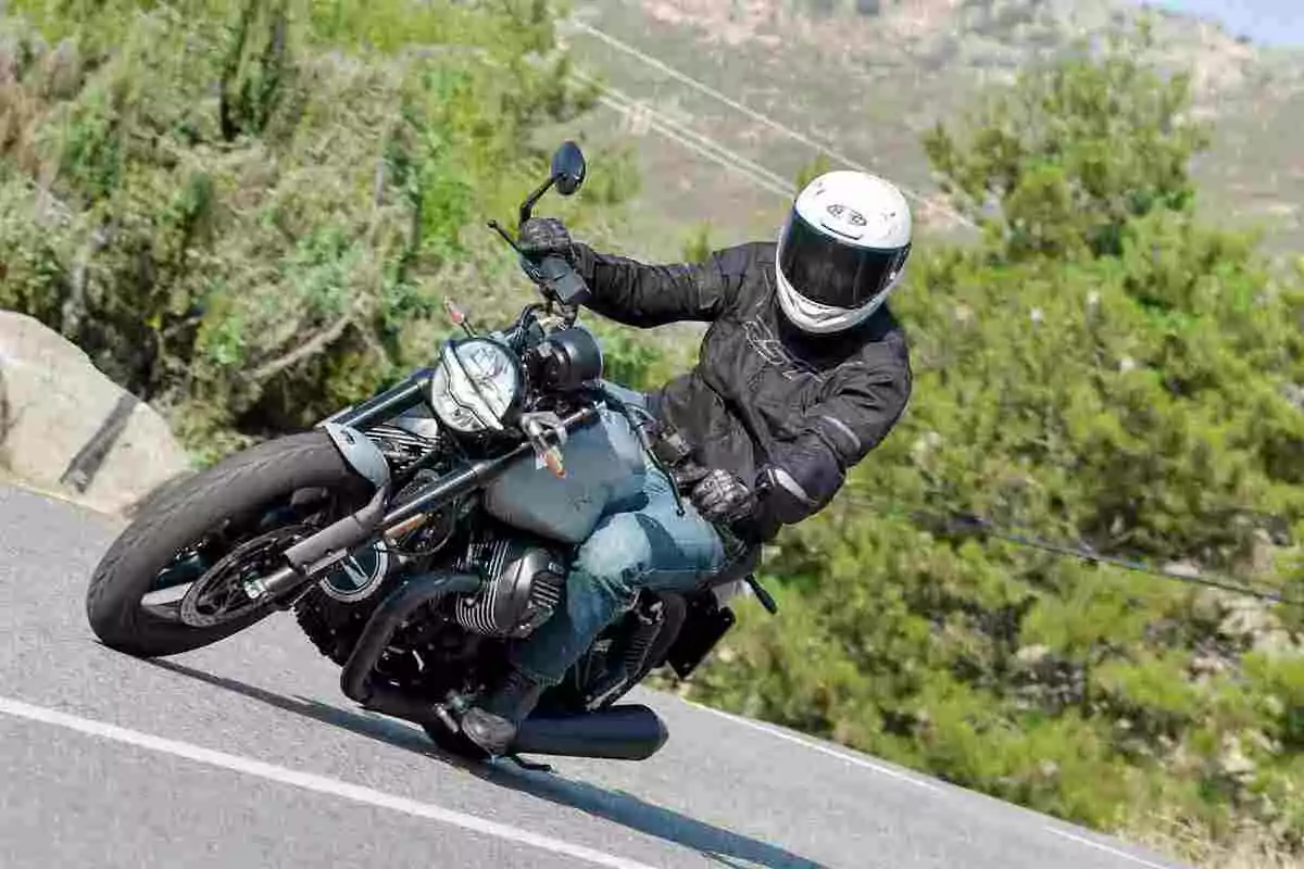 Persona con casco blanco y chaqueta negra conduciendo una motocicleta en una carretera con vegetación al fondo Persona con casco blanco y chaqueta negra conduciendo una motocicleta en una carretera con vegetación al fondo