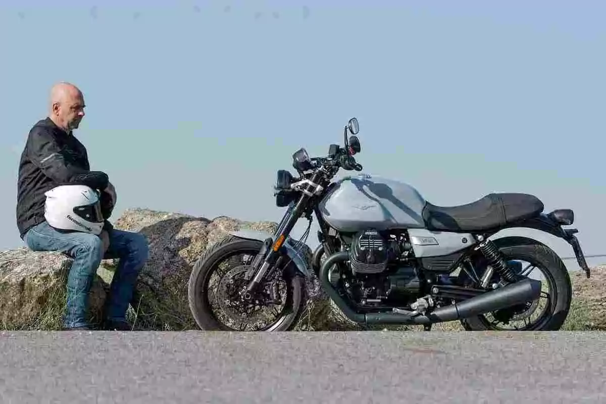 Hombre sentado en una roca sosteniendo un casco blanco mientras observa una motocicleta estacionada en un camino asfaltado bajo un cielo despejado Hombre sentado en una roca sosteniendo un casco blanco mientras observa una motocicleta estacionada en un camino asfaltado bajo un cielo despejado