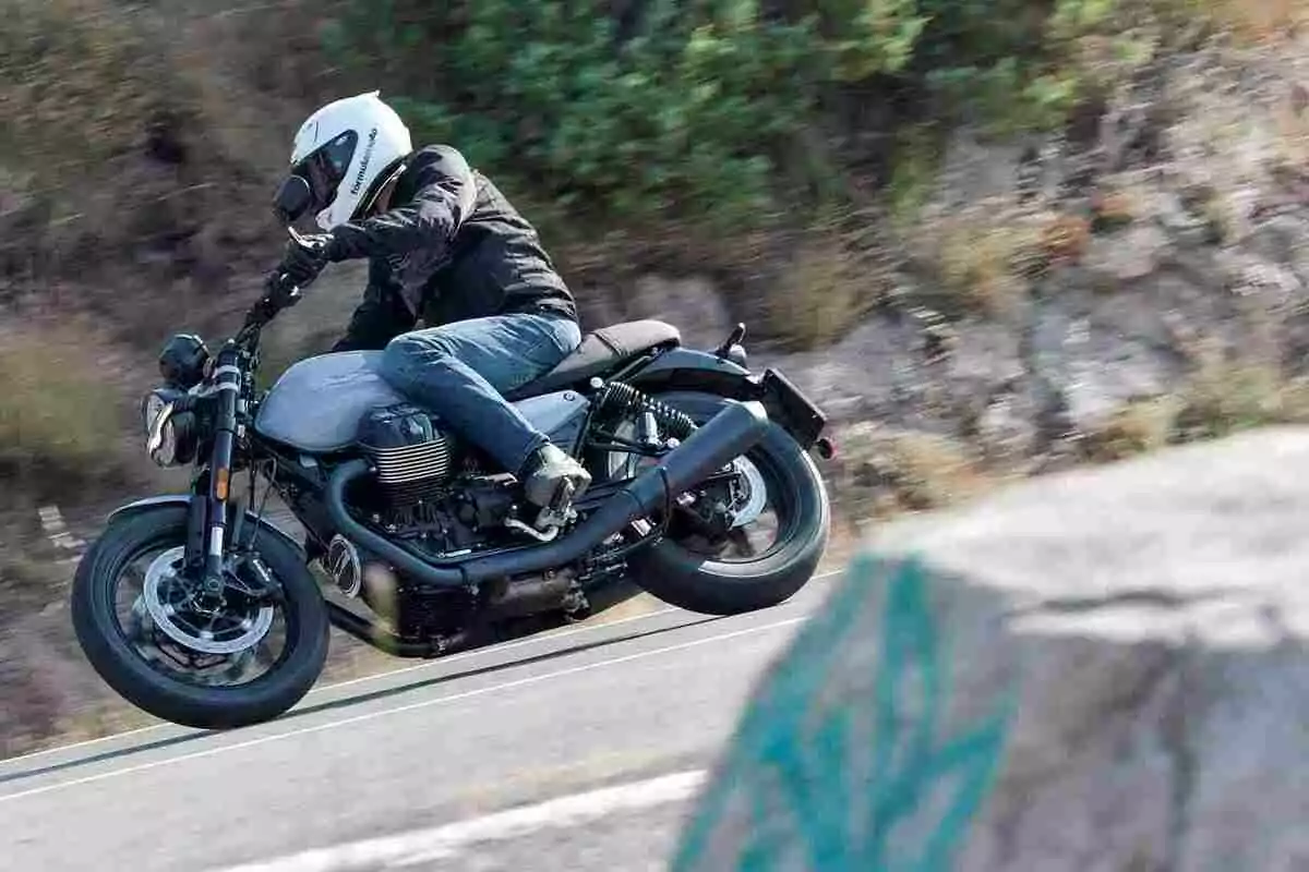 Persona con casco blanco y chaqueta negra conduciendo una motocicleta gris en una carretera con curvas y vegetación al fondo Persona con casco blanco y chaqueta negra conduciendo una motocicleta gris en una carretera con curvas y vegetación al fondo
