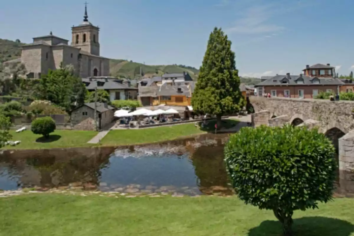 Vista de un pueblo con iglesia de piedra, casas tradicionales, un puente antiguo y un río rodeado de césped y árboles en un día soleado Vista de un pueblo con iglesia de piedra, casas tradicionales, un puente antiguo y un río rodeado de césped y árboles en un día soleado
