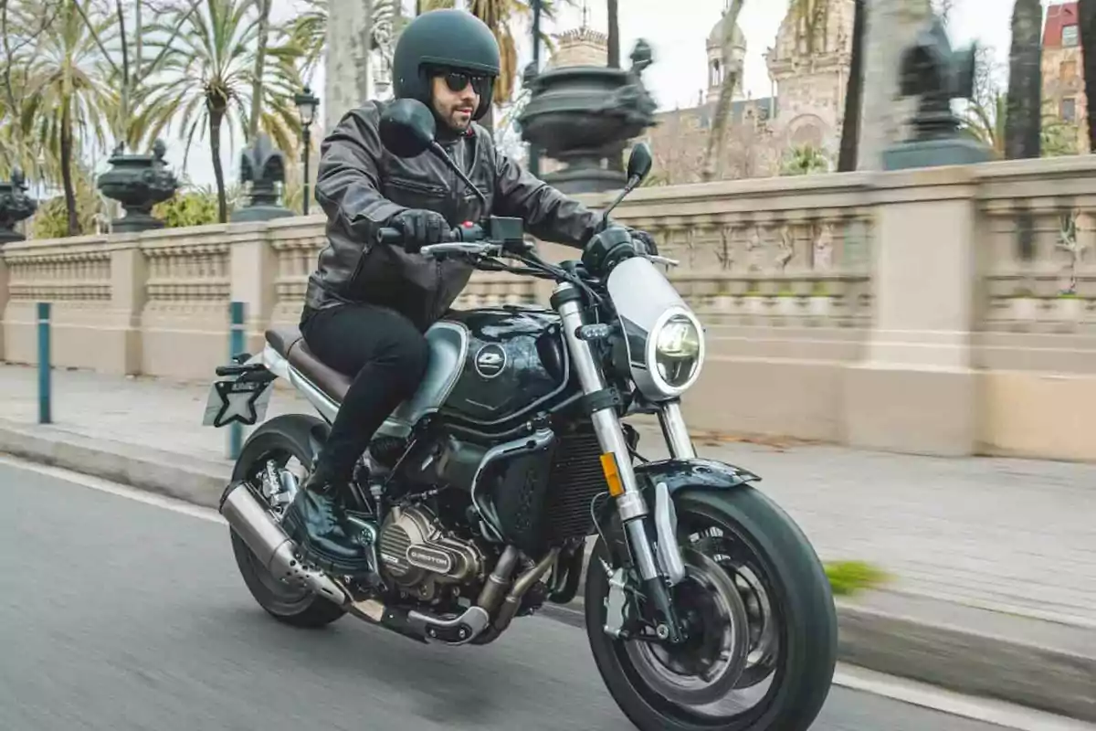 Hombre conduciendo una motocicleta negra por una calle urbana con palmeras y edificios al fondo Hombre conduciendo una motocicleta negra por una calle urbana con palmeras y edificios al fondo