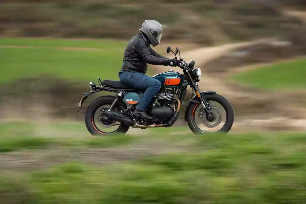 Persona con casco y chaqueta negra conduciendo una motocicleta en movimiento sobre un camino rodeado de vegetación Persona con casco y chaqueta negra conduciendo una motocicleta en movimiento sobre un camino rodeado de vegetación