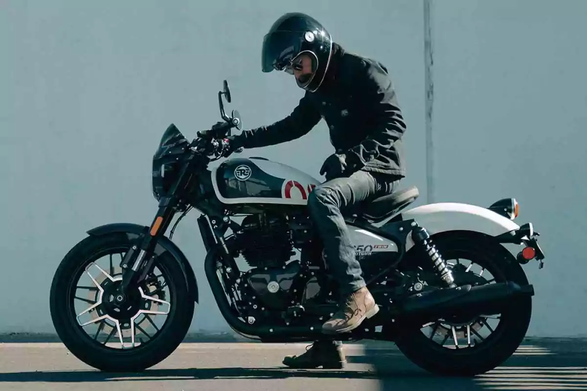 Persona con casco y chaqueta negra sentada sobre una motocicleta blanca junto a una pared clara Persona con casco y chaqueta negra sentada sobre una motocicleta blanca junto a una pared clara