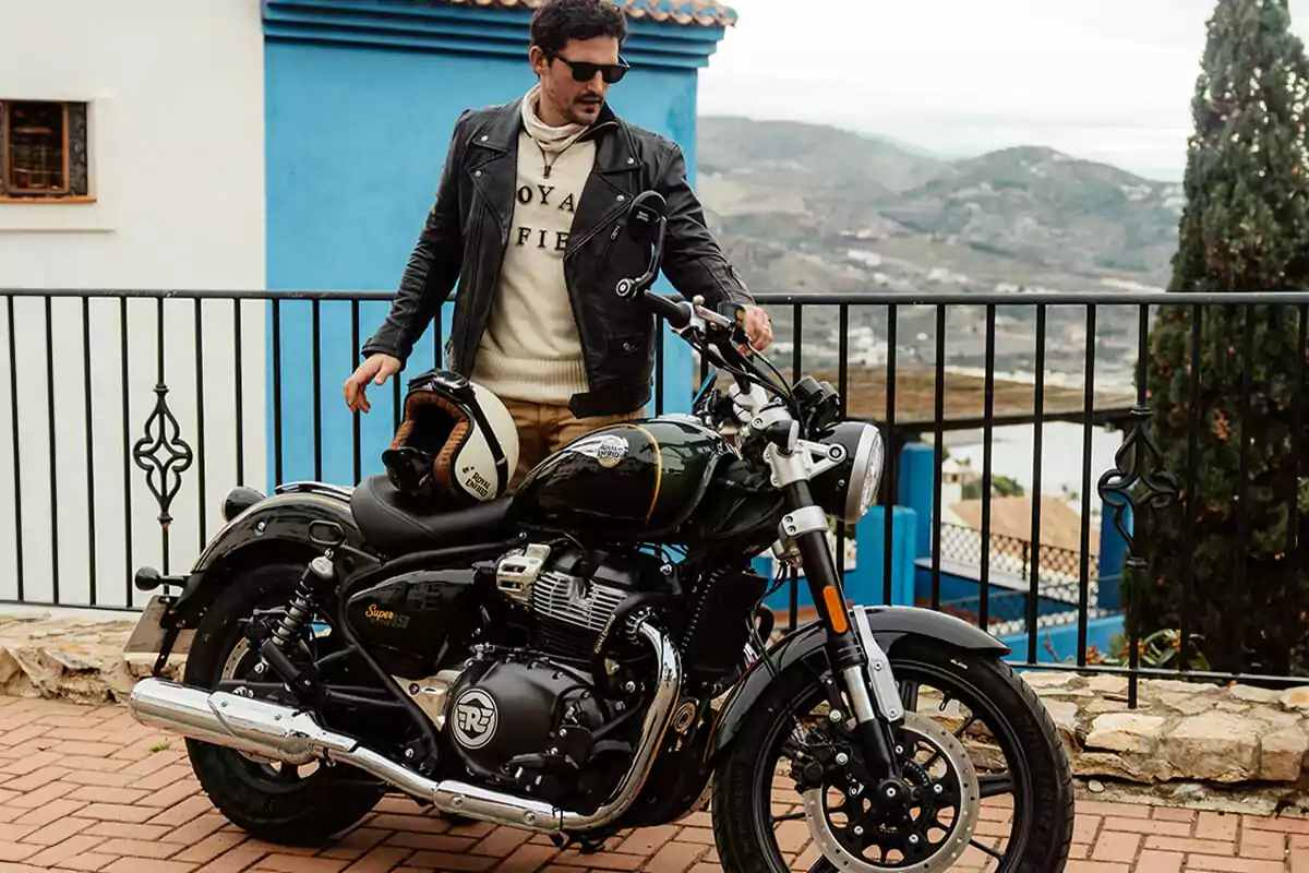Hombre con chaqueta de cuero y gafas de sol junto a una motocicleta negra estacionada en una terraza con paisaje de montaña al fondo Hombre con chaqueta de cuero y gafas de sol junto a una motocicleta negra estacionada en una terraza con paisaje de montaña al fondo