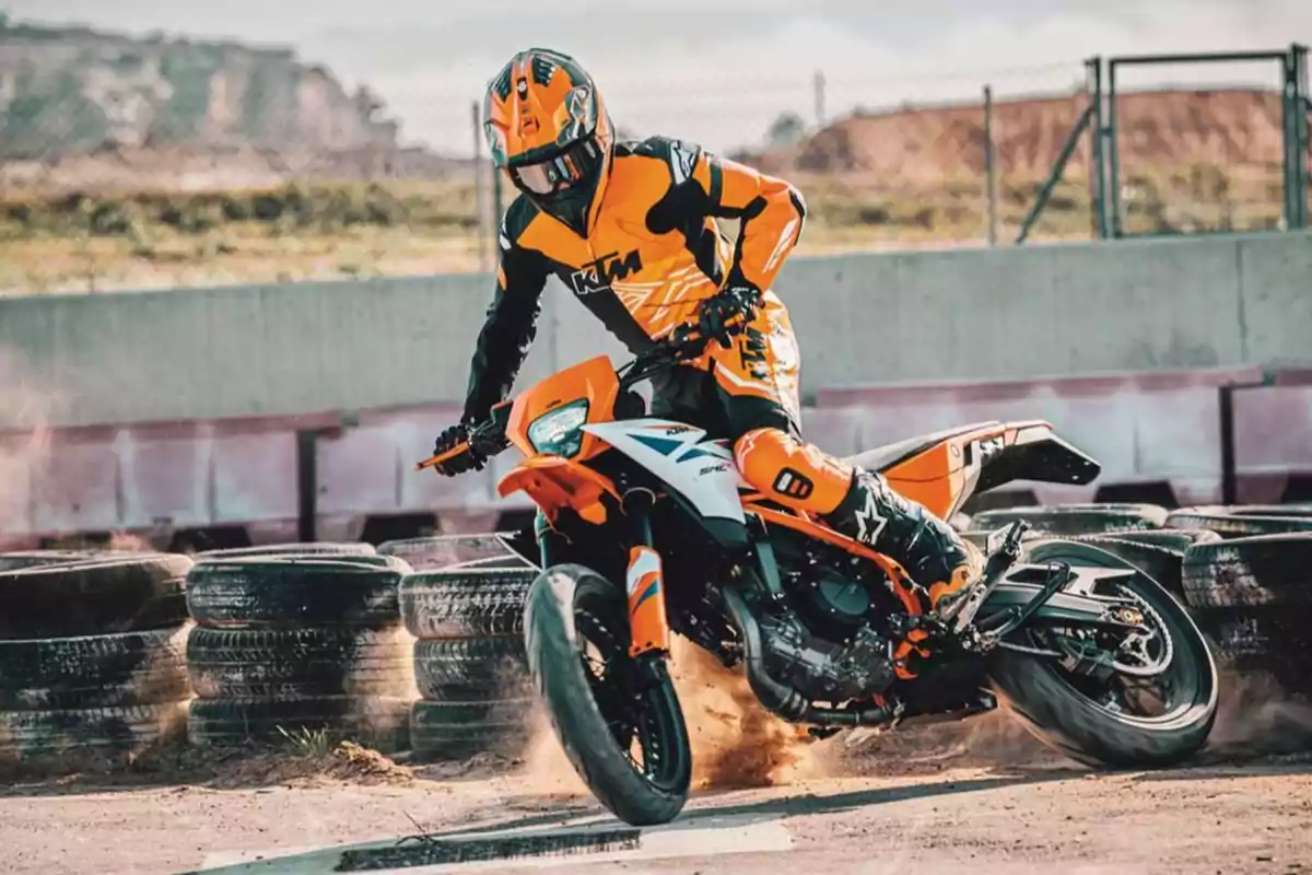 Piloto con traje y casco naranja conduciendo una motocicleta KTM en una pista de tierra junto a una barrera de llantas Piloto con traje y casco naranja conduciendo una motocicleta KTM en una pista de tierra junto a una barrera de llantas