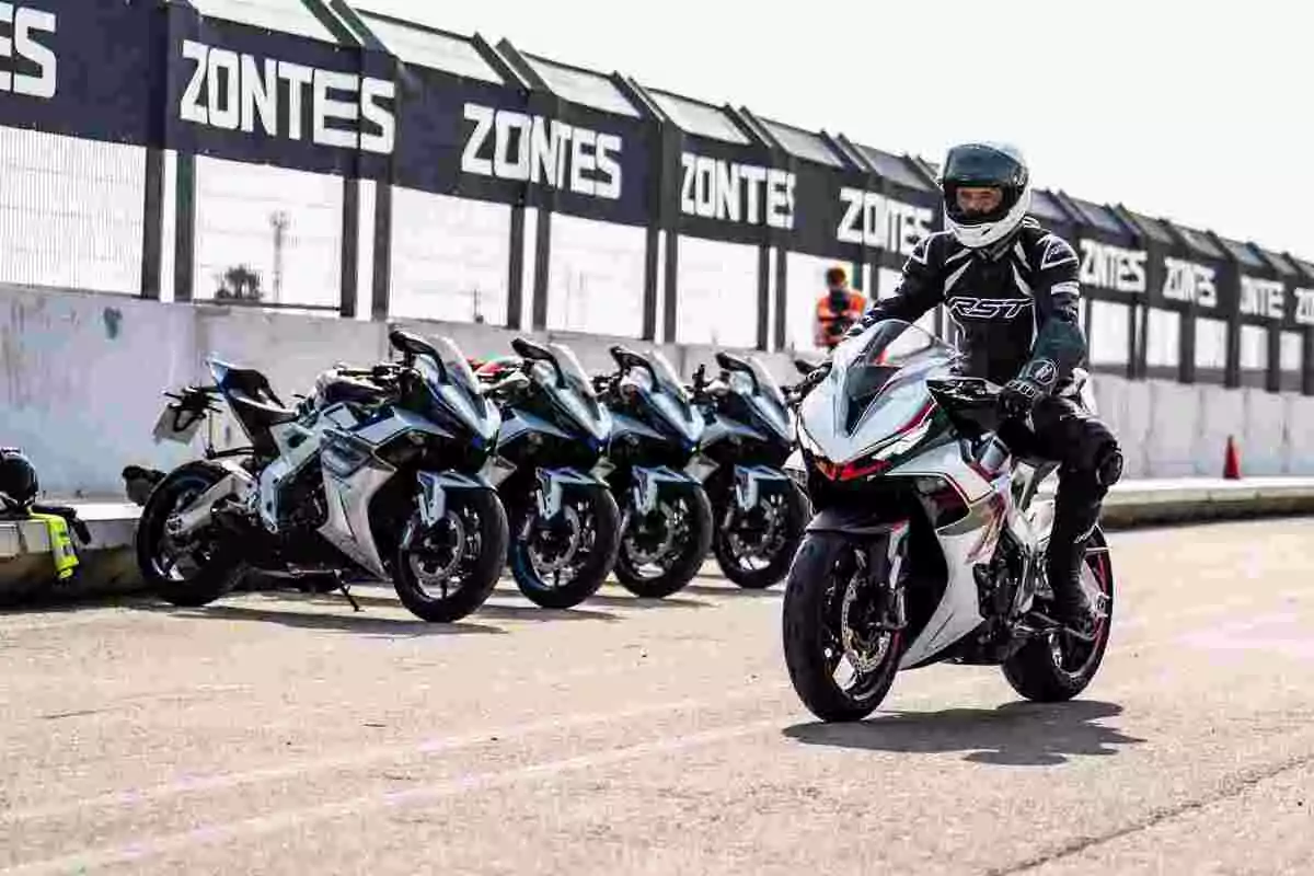 Pilot amb equip de protecció conduint una motocicleta esportiva blanca en un circuit, mentre diverses motos similars estan alineades al fons al costat d’una tanca publicitària de Zontes. Pilot amb equip de protecció conduint una motocicleta esportiva blanca en un circuit, mentre diverses motos similars estan alineades al fons al costat d’una tanca publicitària de Zontes.