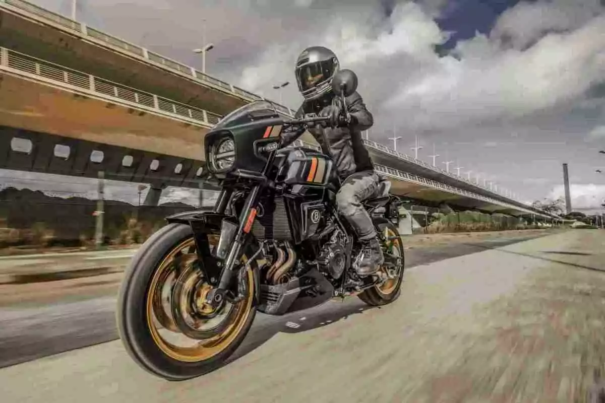 Persona conduciendo una motocicleta negra con detalles naranjas y dorados en una vía urbana junto a un puente moderno bajo un cielo parcialmente nublado Persona conduciendo una motocicleta negra con detalles naranjas y dorados en una vía urbana junto a un puente moderno bajo un cielo parcialmente nublado