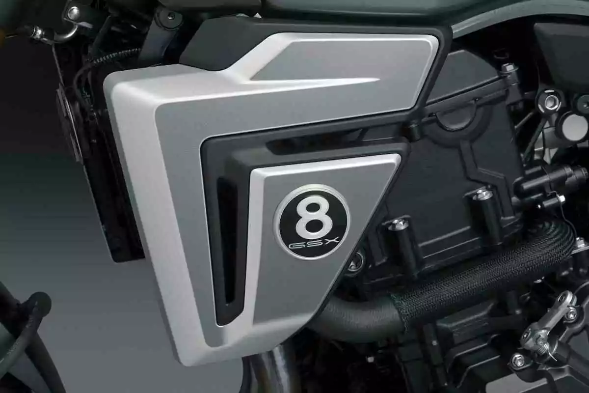 Detalle del motor de una motocicleta con una cubierta plateada que tiene el número 8 y las letras GSX en un círculo negro Detalle del motor de una motocicleta con una cubierta plateada que tiene el número 8 y las letras GSX en un círculo negro