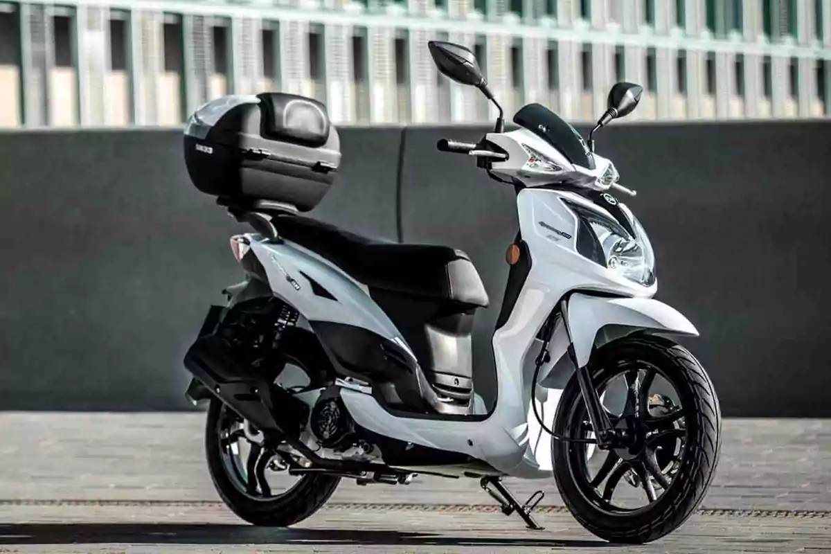 Motocicleta tipus scooter blanca estacionada sobre el paviment amb un bagul negre a la part del darrere i fons urbà desenfocat Motocicleta tipus scooter blanca estacionada sobre el paviment amb un bagul negre a la part del darrere i fons urbà desenfocat