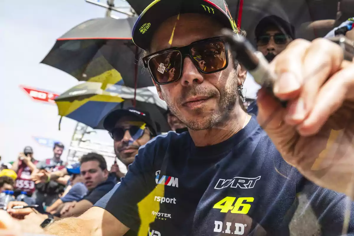 Hombre con gafas de sol y gorra firmando un autógrafo mientras varias personas lo observan en un evento al aire libre Hombre con gafas de sol y gorra firmando un autógrafo mientras varias personas lo observan en un evento al aire libre