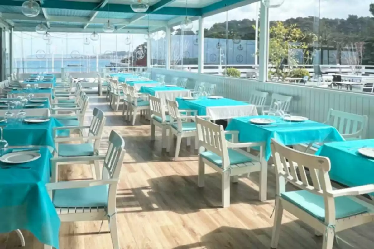 Terraza de restaurante con mesas y sillas blancas, manteles turquesa y vista al mar Terraza de restaurante con mesas y sillas blancas, manteles turquesa y vista al mar