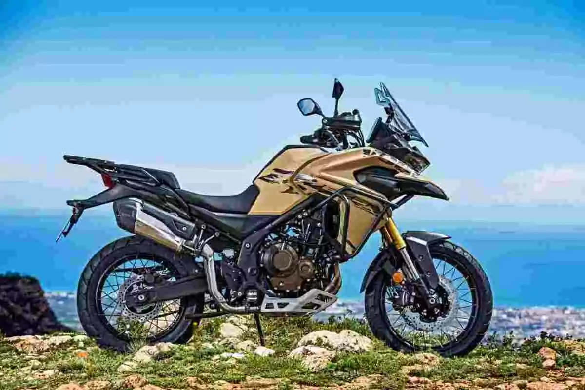 Motocicleta tipo adventure color dorado estacionada sobre un terreno rocoso con el mar y el cielo azul de fondo Motocicleta tipo adventure color dorado estacionada sobre un terreno rocoso con el mar y el cielo azul de fondo