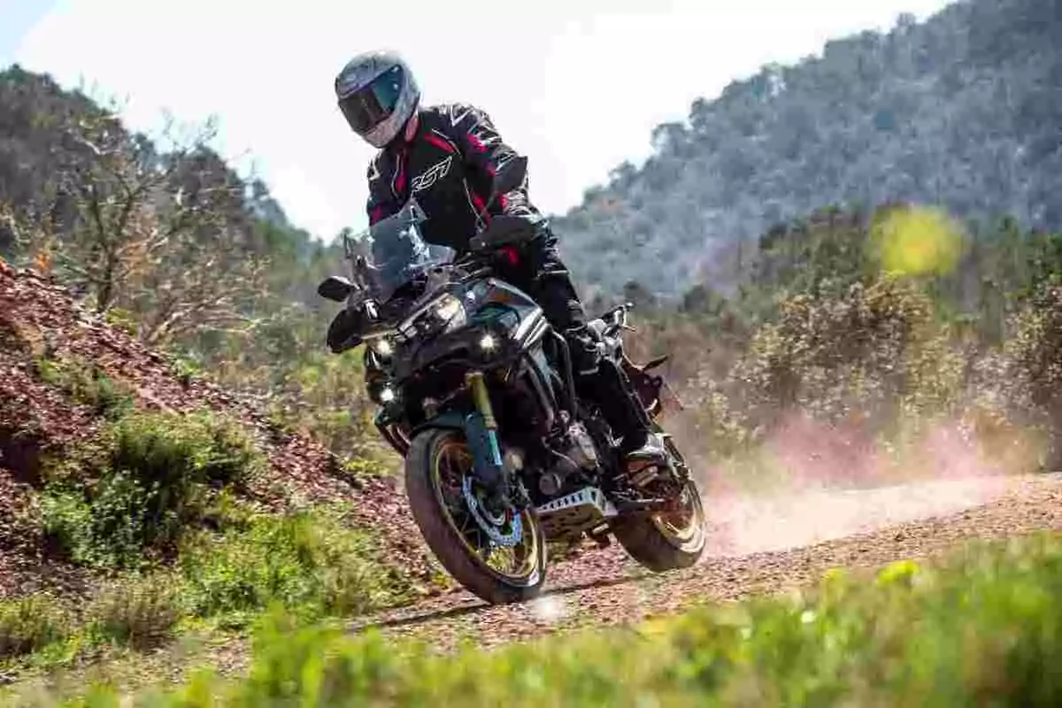 Person riding an adventure motorcycle on a dirt road surrounded by vegetation and mountains in the background
