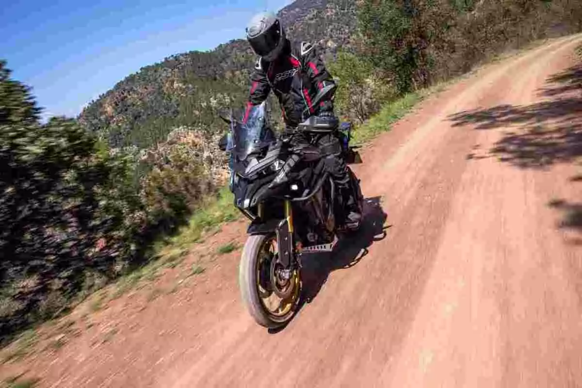 Persona conduciendo una motocicleta deportiva por un camino de tierra rodeado de vegetación y montañas bajo un cielo despejado Persona conduciendo una motocicleta deportiva por un camino de tierra rodeado de vegetación y montañas bajo un cielo despejado