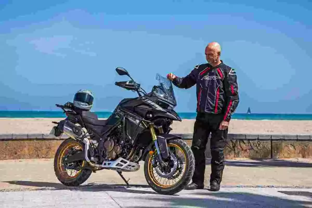 Hombre con equipo de motociclista de pie junto a una moto negra en la playa con el mar de fondo Hombre con equipo de motociclista de pie junto a una moto negra en la playa con el mar de fondo