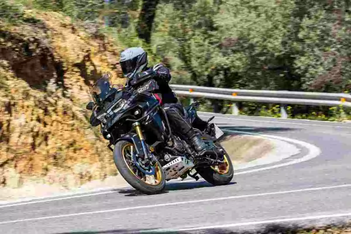Persona con casco y equipo de protección conduciendo una motocicleta deportiva en una curva de carretera rodeada de vegetación Persona con casco y equipo de protección conduciendo una motocicleta deportiva en una curva de carretera rodeada de vegetación
