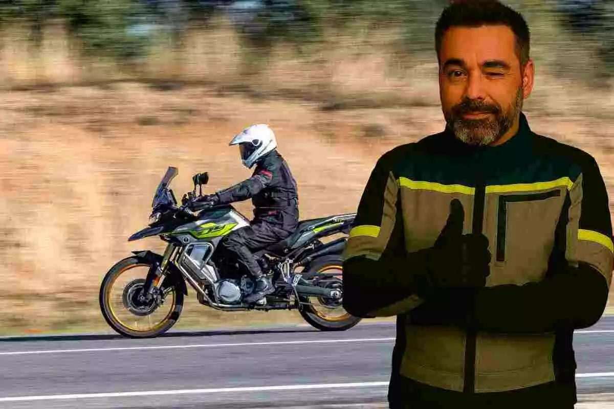 Home amb jaqueta de motorista fent un gest d’aprovació mentre al fons una persona condueix una moto per la carretera.