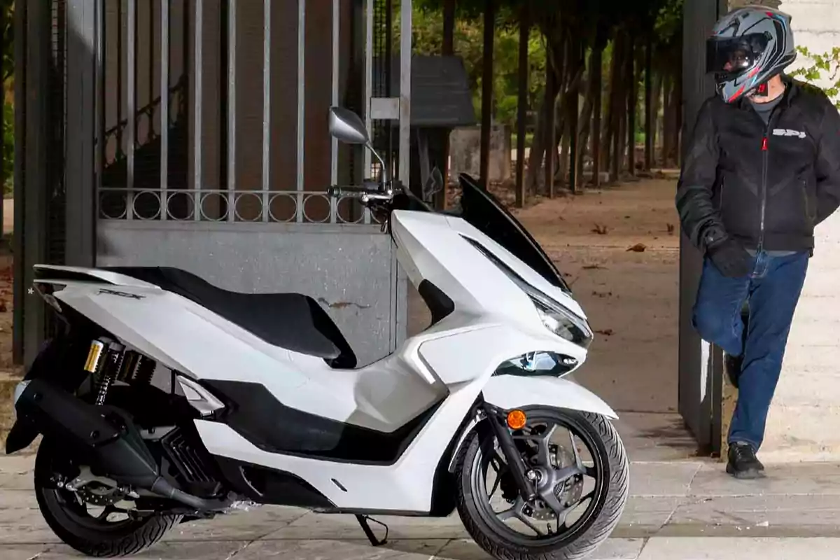 Motocicleta blanca tipo scooter estacionada junto a una reja con una persona usando casco y chaqueta de motociclista apoyada en la entrada Motocicleta blanca tipo scooter estacionada junto a una reja con una persona usando casco y chaqueta de motociclista apoyada en la entrada