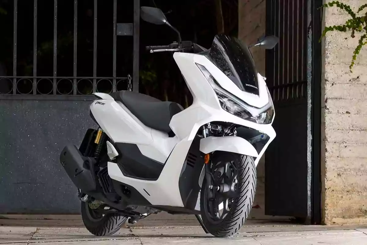 Motocicleta tipo scooter color blanco estacionada frente a una reja de metal y una pared de ladrillo Motocicleta tipo scooter color blanco estacionada frente a una reja de metal y una pared de ladrillo