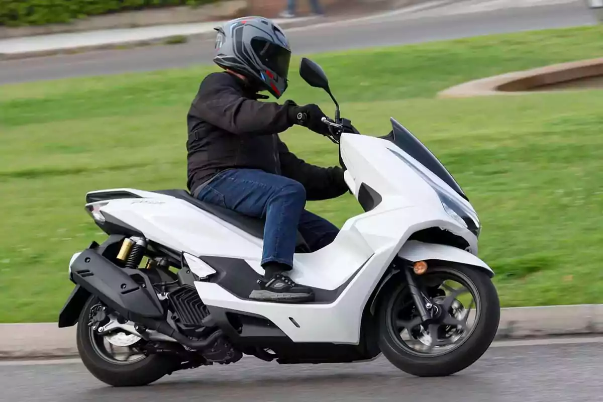 Persona conduciendo una motocicleta tipo scooter blanca en una calle junto a un área verde