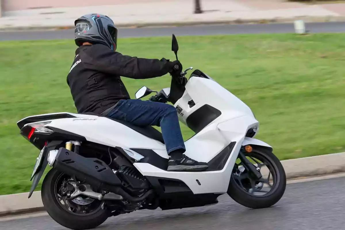 Persona conduciendo una motocicleta tipo scooter blanca en una calle junto a un área verde Persona conduciendo una motocicleta tipo scooter blanca en una calle junto a un área verde