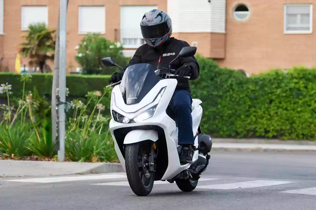 Persona conduciendo una moto tipo scooter blanca en una calle urbana con vegetación y edificios al fondo Persona conduciendo una moto tipo scooter blanca en una calle urbana con vegetación y edificios al fondo