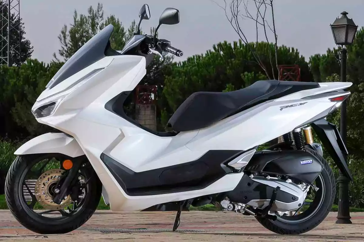 Motocicleta tipo scooter de color blanco estacionada en un entorno al aire libre con árboles y farolas al fondo Motocicleta tipo scooter de color blanco estacionada en un entorno al aire libre con árboles y farolas al fondo