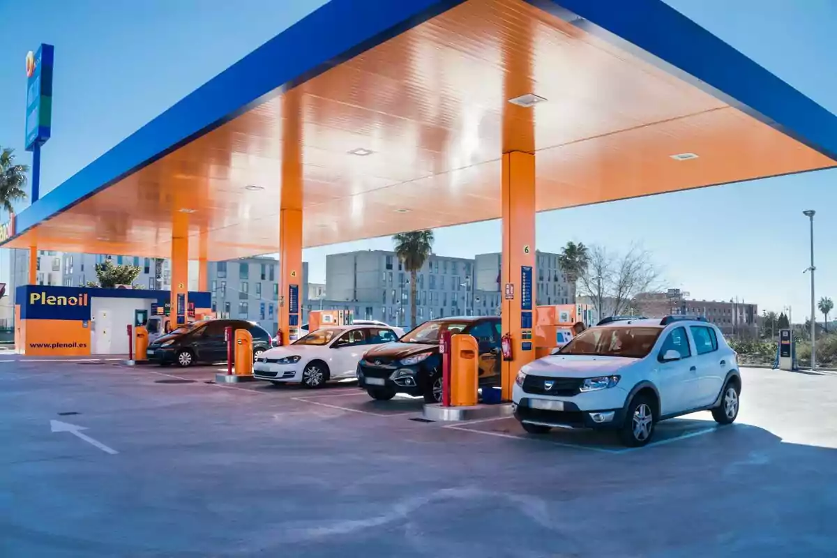 Una estación de servicio con varios coches estacionados bajo un techo naranja y azul, con surtidores de combustible y un edificio de fondo. Una estación de servicio con varios coches estacionados bajo un techo naranja y azul, con surtidores de combustible y un edificio de fondo.