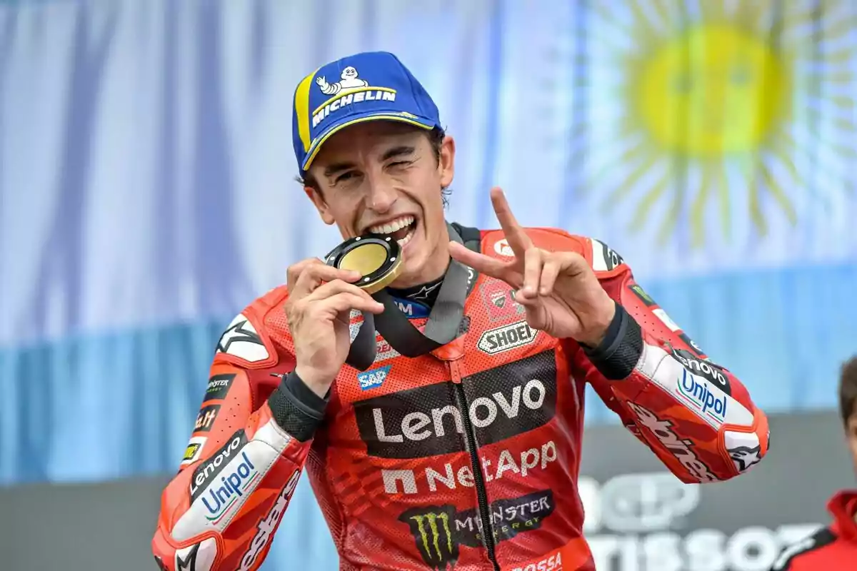 Un motociclista sonriente con uniforme rojo y gorra azul sostiene una medalla frente a una bandera. Un motociclista sonriente con uniforme rojo y gorra azul sostiene una medalla frente a una bandera.