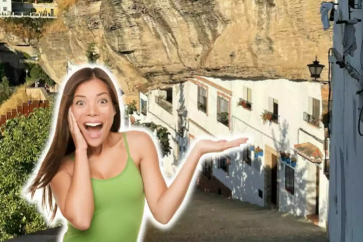 Una mujer con una camiseta verde muestra una expresión de sorpresa mientras señala con la mano un conjunto de casas blancas construidas bajo una gran roca. Una mujer con una camiseta verde muestra una expresión de sorpresa mientras señala con la mano un conjunto de casas blancas construidas bajo una gran roca.
